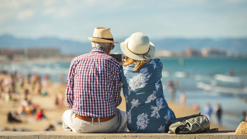 An older couple sitting on a wall at a beach.