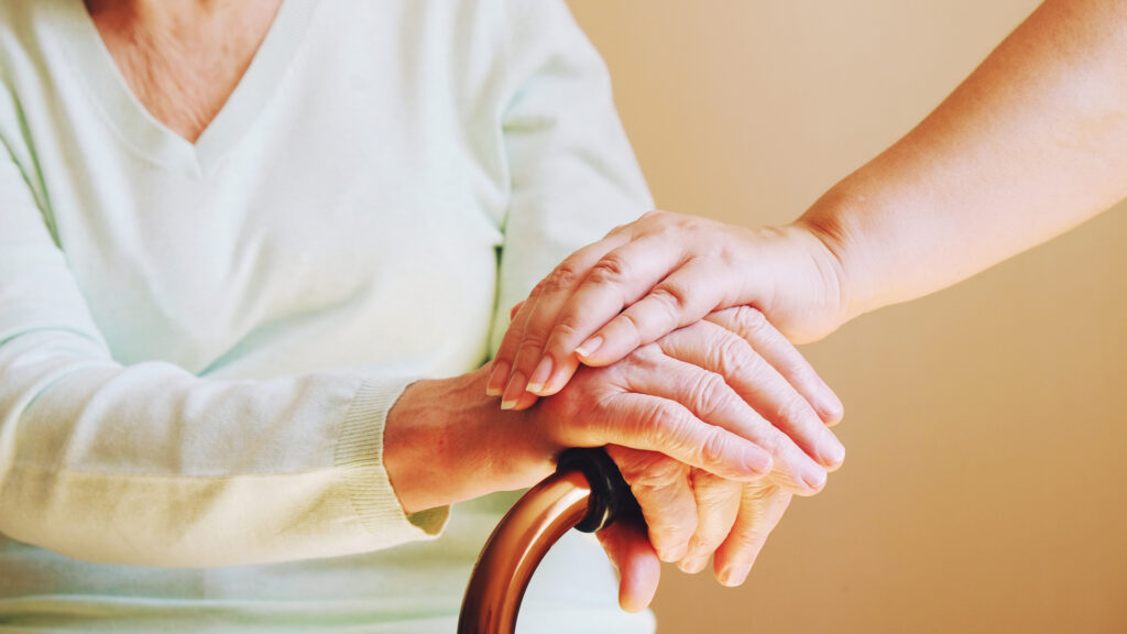 Close-up of someone holding hand the hand of an older woman with a walking aid.